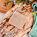 Bar of soap on a wooden surface with a plant and towel in the background
